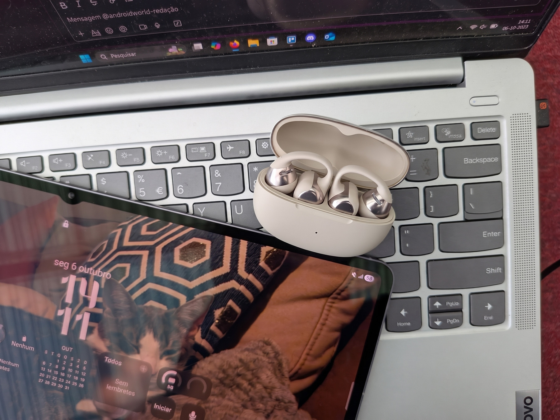 Auriculares sem fios na caixa sobre o teclado de um portátil prateado, com um tablet em primeiro plano exibindo um gato no ecrã de bloqueio.
